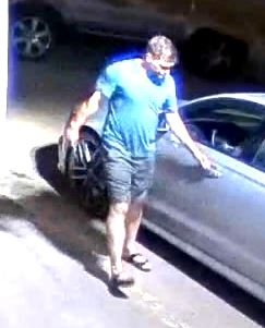 From an elevated camera, the man walking on the pavement beside a dark parked vehicle.